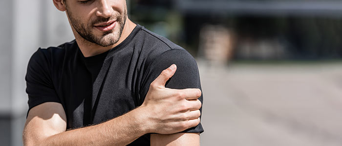 man in tshirt holding his shoulder in pain, he is outside by a wall